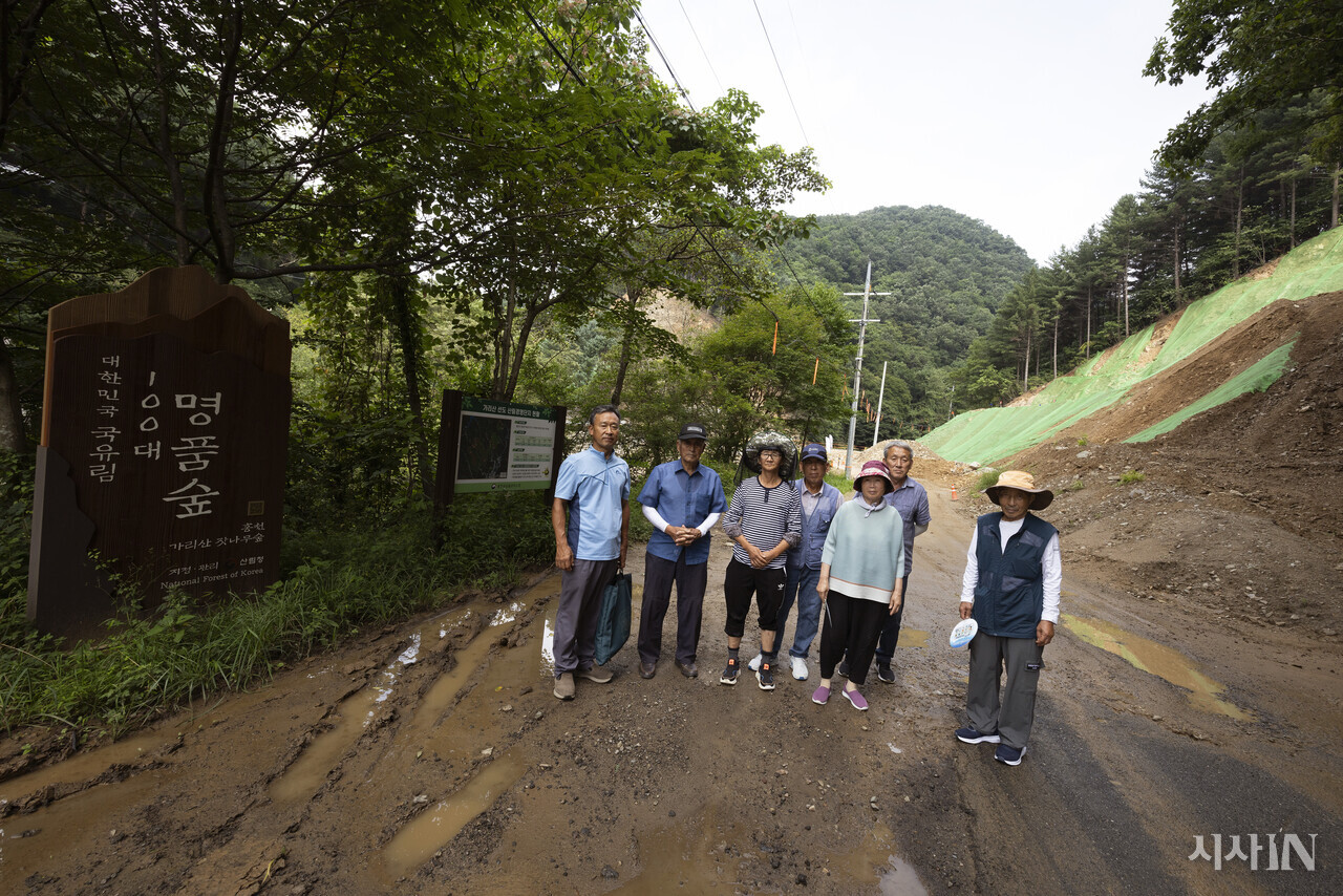 강원도 홍천군 풍천리 마을 주민인 이창후(61·왼쪽부터) 박시현(83) 변진호(56) 박영식(74) 서도화(80) 박명식(68) 김용식씨(82)가 이설도로 공사가 한창인 대한민국 100대 명품숲 간판 앞에서 사진을 찍고 있다. 홍천군 풍천리의 잣나무 숲은 2017년 ‘명품숲 10선’에, 2023년 ‘대한민국 100대 명품숲’에 지정되었다. ⓒ시사IN 이명익